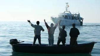 تظهر في الصورة مجموعة من الأشخاص يقفون في قارب صغير في المياه الهادئة، ويبدو أنهم يلوحون بأيديهم نحو قارب أكبر يقترب منهم. المشهد يعكس نشاطًا بحريًا، حيث الأجواء هادئة وتظهر السماء والماء بلونٍ صافٍ. الأشخاص يظهرون من الخلف، مما يركز الانتباه على الإيماءات التي يقومون بها.